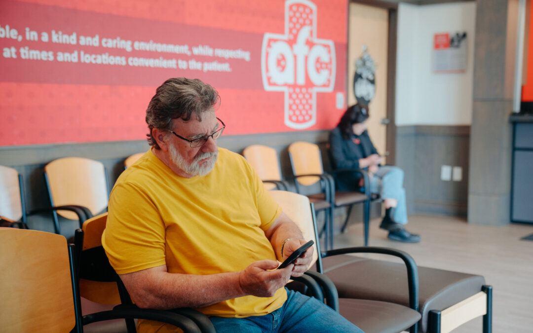 man waiting in reception area of an AFC Urgent Care clinic while looking at his phone while woman in mask also waits in the background.